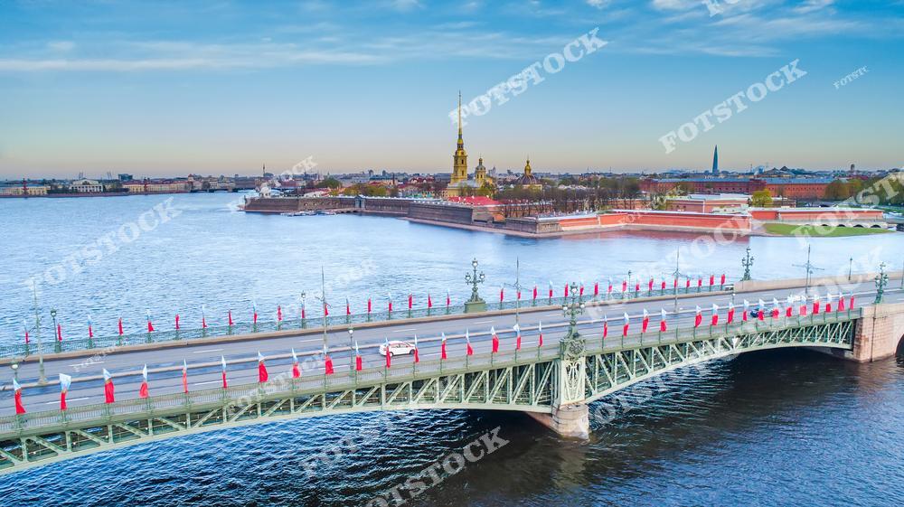 На фотографии изображен Троицкий мост, украшенный праздничными флагами, простирающийся через реку Неву. На заднем плане видна Петропавловская крепость с ее золотыми шпилями, которая является одной из главных достопримечательностей Санкт-Петербурга. Левее виднеются исторические здания Васильевского острова. Река Нева в этот момент отражает свет утреннего неба, создавая живописный вид.
